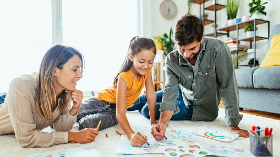 a mother, father, and daughter cheerfully draw together, papers and drawing materials spread across a living room floor