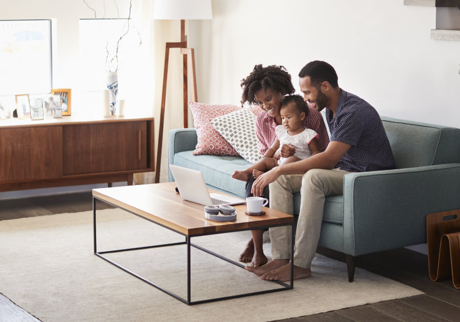 A mom and dad and their baby child sit on a living-room couch together, looking at a laptop computer